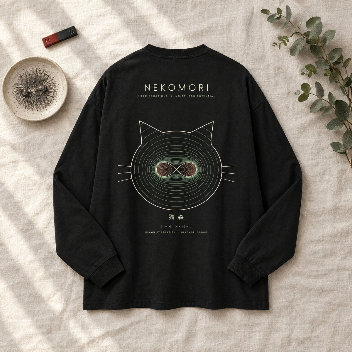 Generated mockup: the black Equipotential long sleeve tee laid back-side up on washed linen beside a bar magnet, iron filings in a ceramic dish and a sprig of eucalyptus.