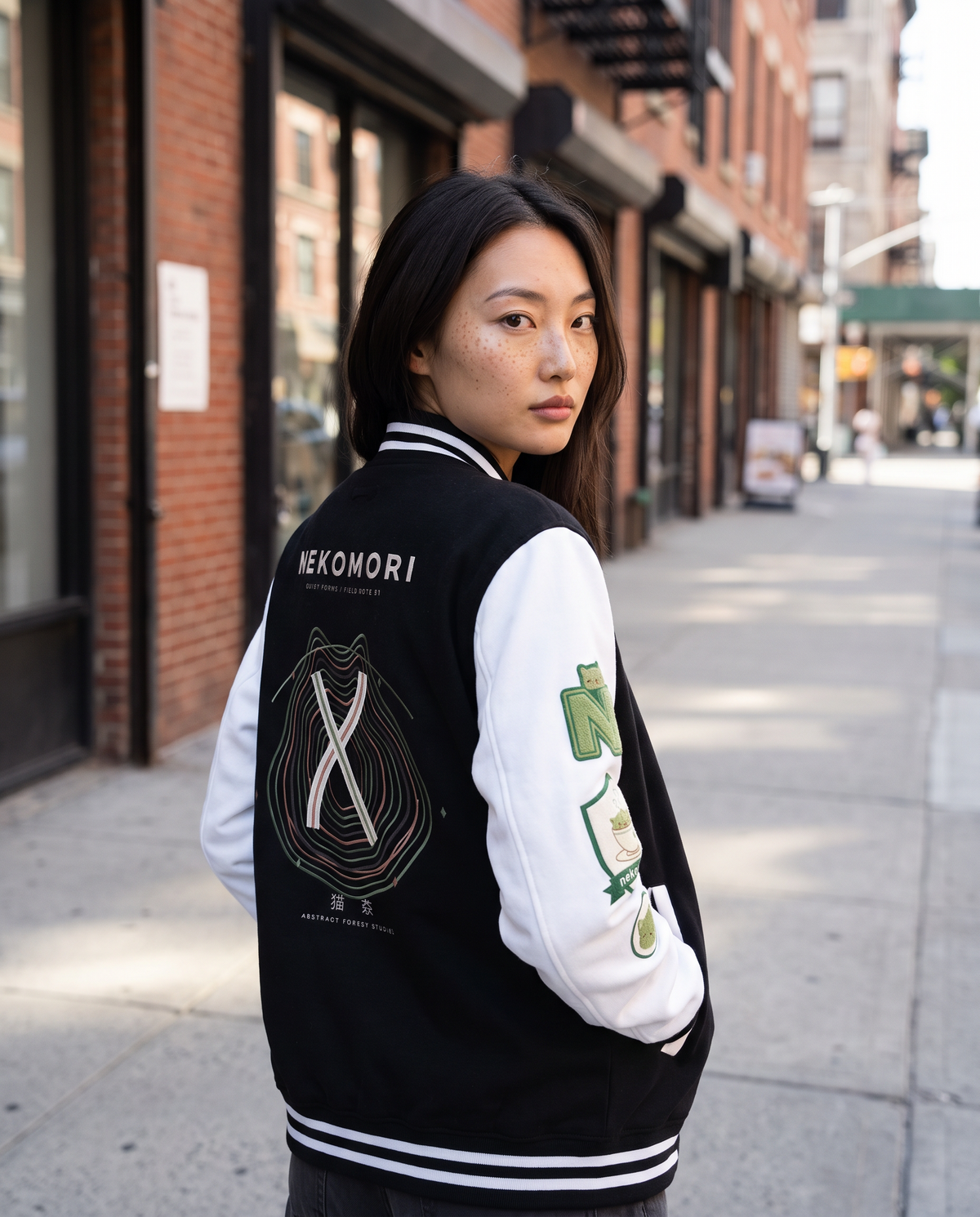 A woman stands on a city sidewalk wearing a black and white varsity jacket with green and white graphics on the sleeve and a large back graphic. The background shows a blurred urban street scene with brick buildings.
